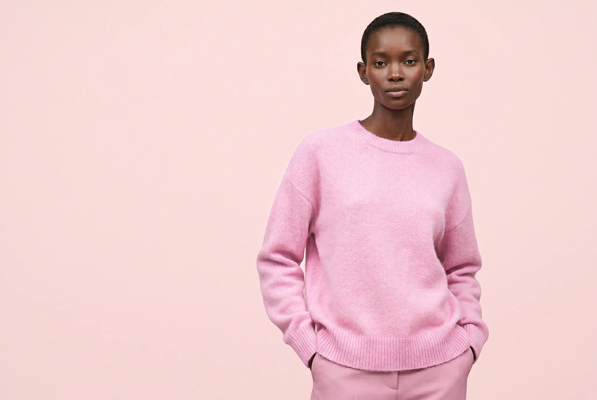 Female model in a pink knit sweater and matching trousers on a pale pink background.